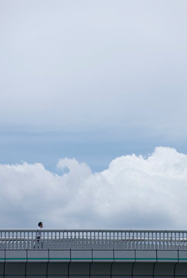 歩道橋を渡る人とその向こうに広がる雲。人は雲から何を学ぶのか