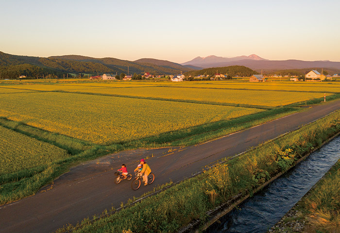 夕日が照らす美しい水田のなかをサイクリング。奥に見えるのは大雪山連峰で、右端のピークが道内最高峰の旭岳（標高2291m）（提供：大塚友記憲さん）