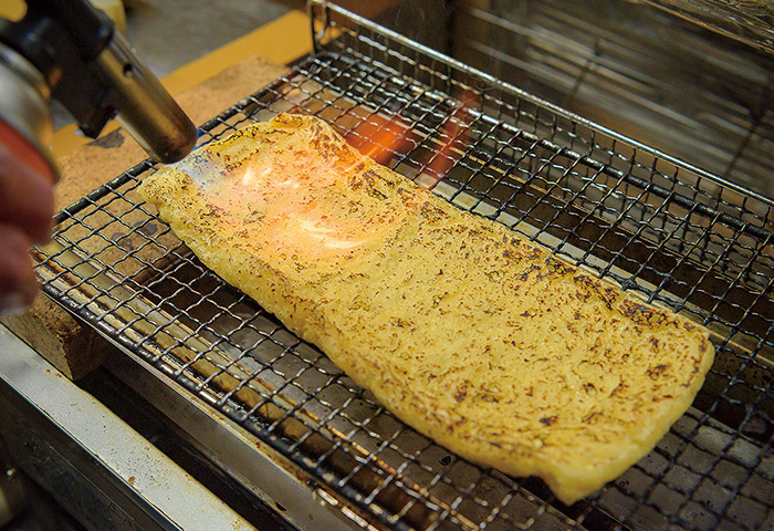 ［２］油揚げはほどよく焦げ目がつくくらいにバーナーで炙ることで、香ばしさとうま味が出る。炙ったら幅2cmほどに切る。1人分の目安は1枚