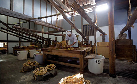 白下糖を白く精製する蜜抜き（押し・研ぎ）と乾燥の作業場。築年不明という古い木造蔵だ。和三盆を水で研ぐ職人・坂東永一さんを囲むように、てこの原理で蜜を絞る押し槽がある